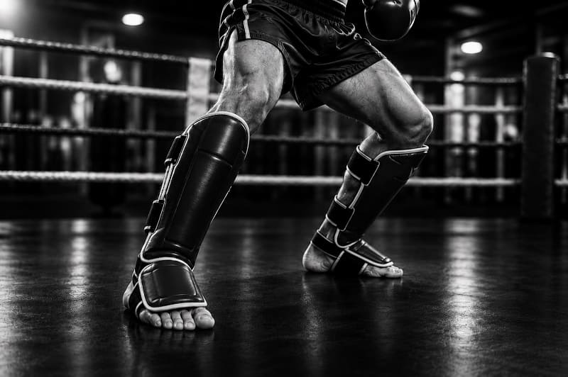 Eine Person steht in einem MMA Cage, im Fokus stehen die Scheinbeinschoner die die Person trägt
